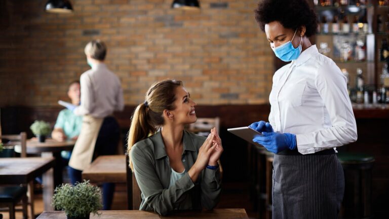 Happy black waitress wearing protective face mask while taking order from customer on a touchpad