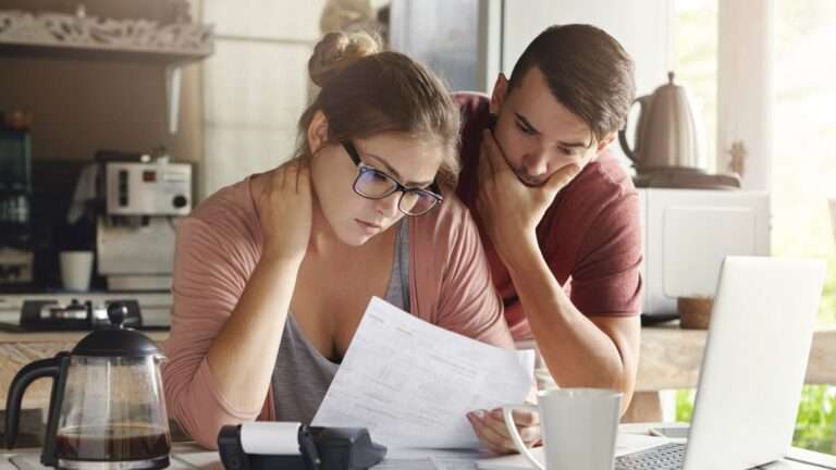 Young caucasian family having debt problems, not able to pay out their loan. female in glasses and brunette man studying paper form bank while managing domestic budget together in kitchen interior